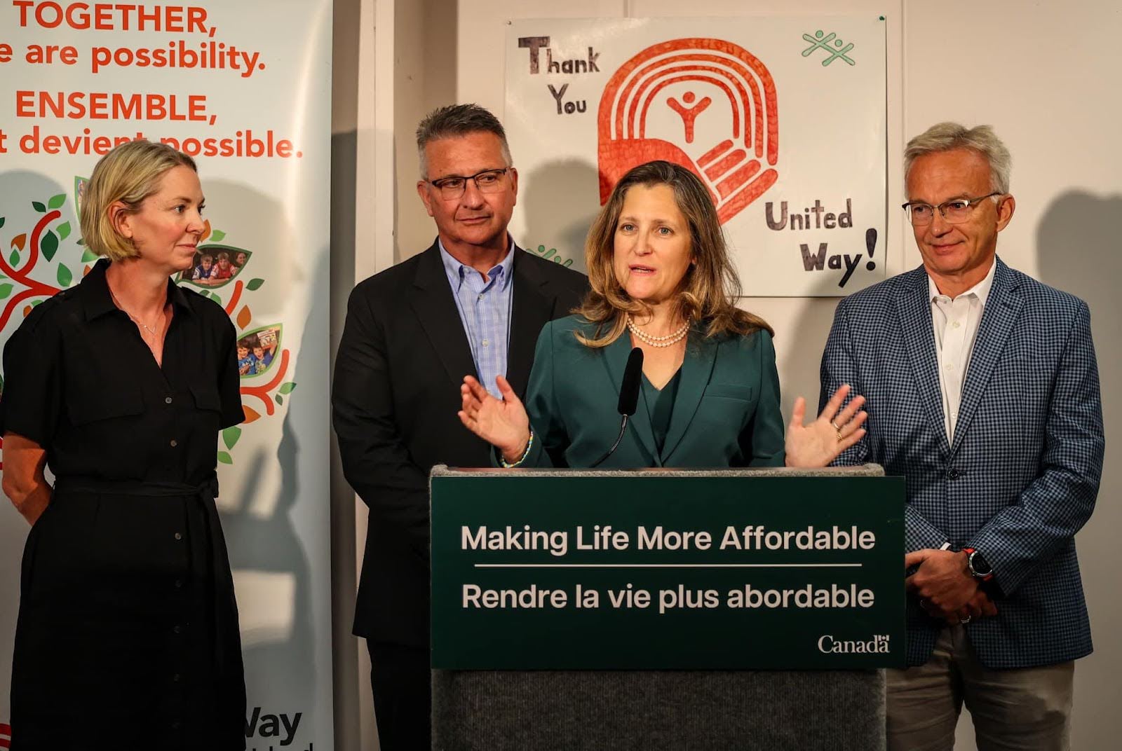 Canadian Finance Minister Chrystia Freeland at the United Way of PEI in Charlottetown, P.E.I. on July 28, 2023. She stands behind a podium with a sign that says "Making Life More Affordable. A woman with shoulder-length blonde hair stands to the left, a man with grey hair wearing a blue shirt and black suit jacket is behind her to the left, and a man with white hair wearing a white shirt and blue plaid suit jacket and grey pants is to the right.