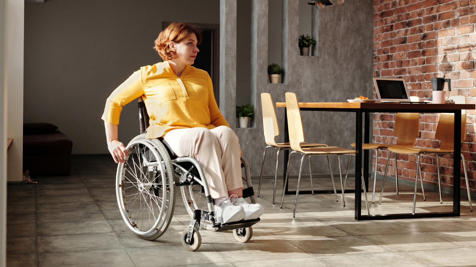 A woman with curly red hair and wearing a yellow shirt, tan pants and white shoes. She is rolling in a wheelchair in an office environment with a brick wall background.