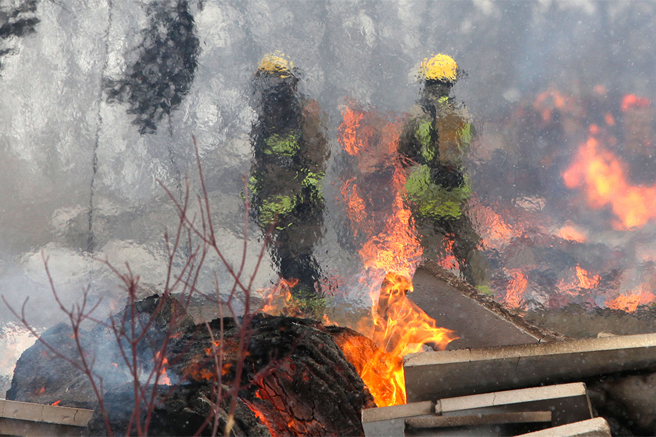 Two people in firefighter gear stand behind orange flames and debris.