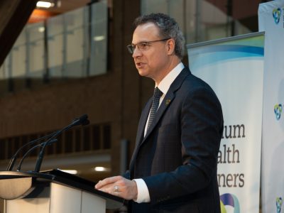 A man in a suit stands a podium in a large room with windows.