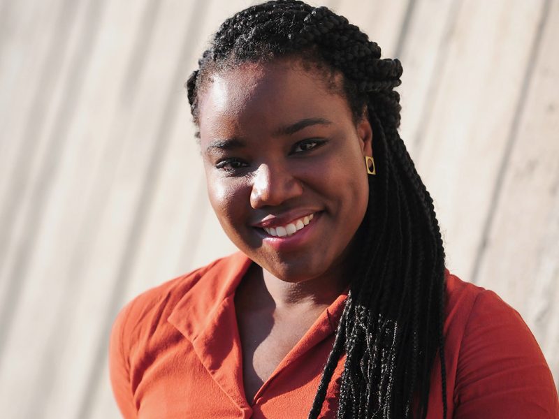 A Black woman in an orange shirt stands in-front of a concrete wall.