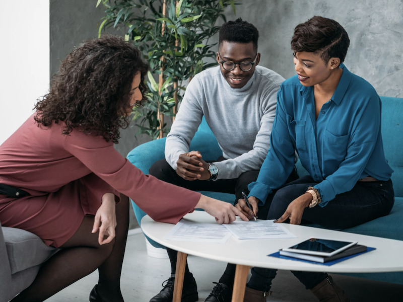 Three young, Black people wearing business casual sit around a low table looking at financial documents.