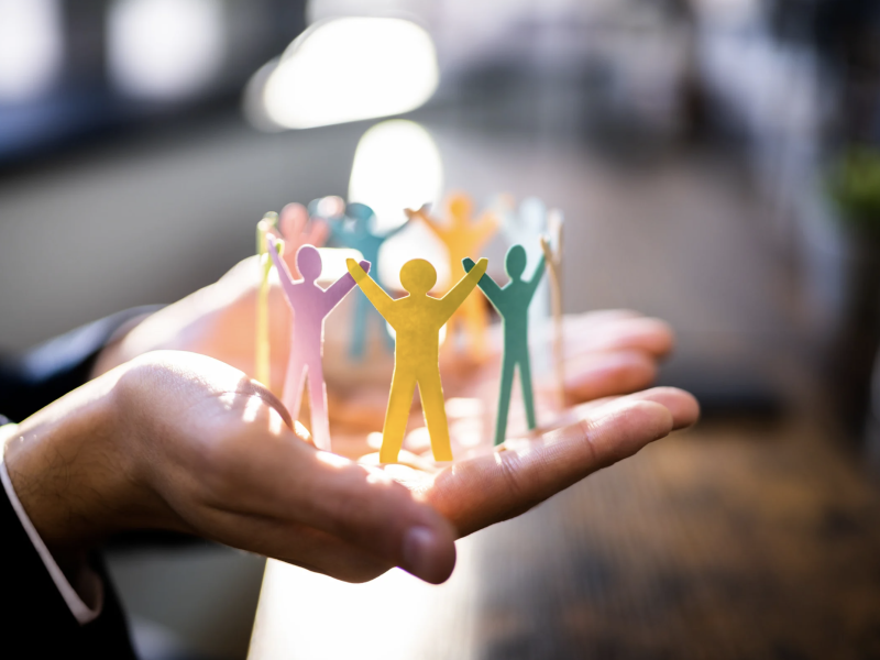 Hands hold multicoloured paper cutouts of people in sunlight.
