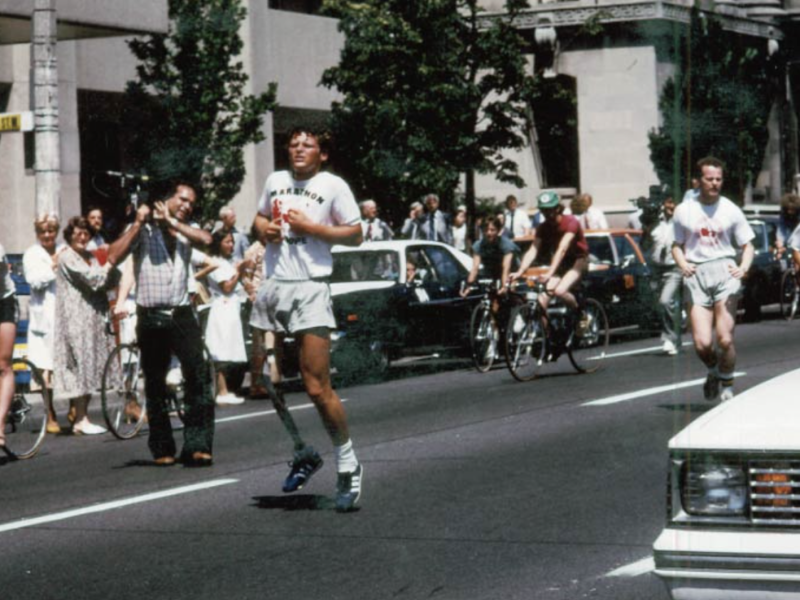 Terry Fox, young man with curly blond hair and a prosthetic leg, runs down a street in the year 1980 while crowds of people cheer him on from the sidewalks.