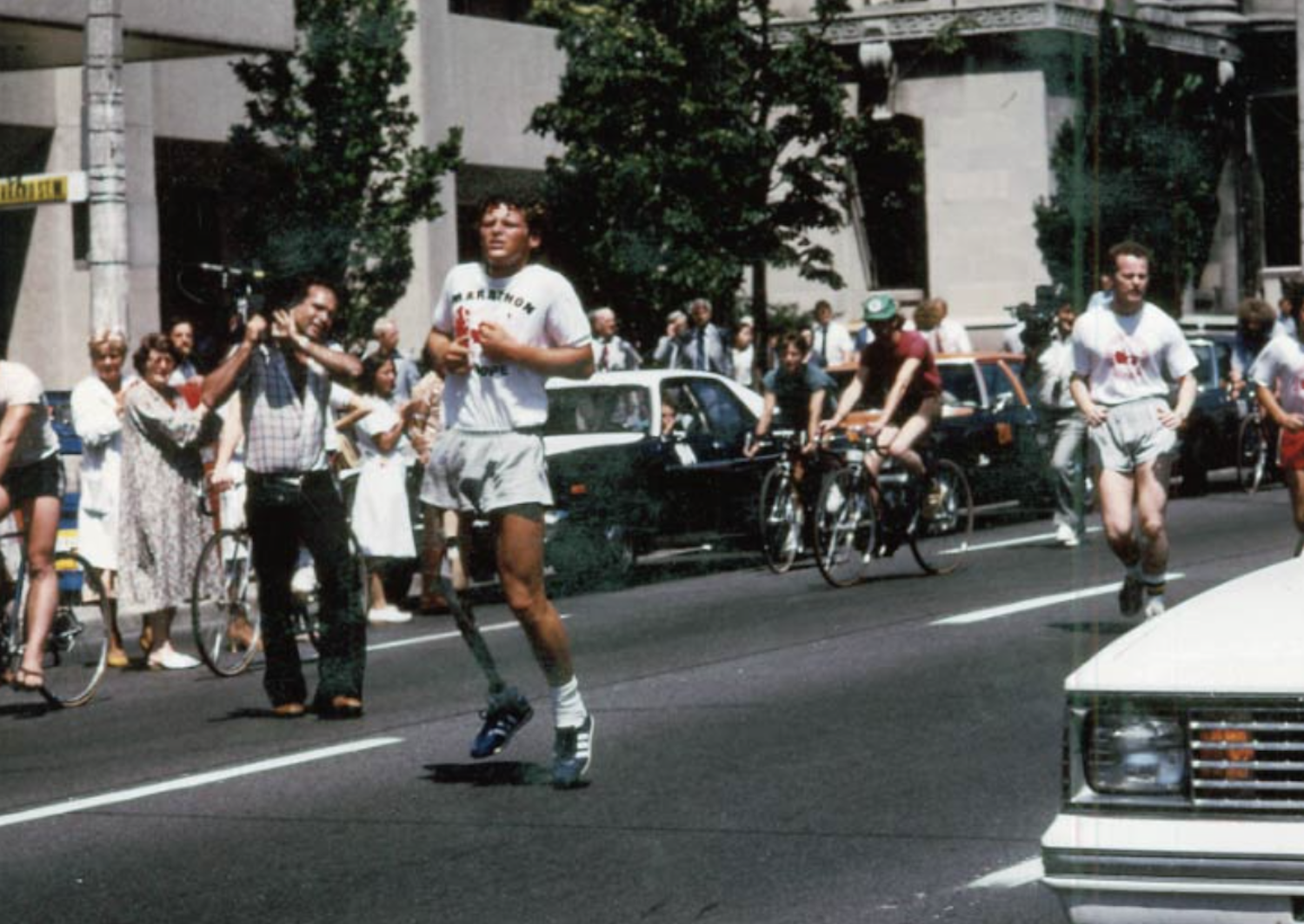 Terry Fox, young man with curly blond hair and a prosthetic leg, runs down a street in the year 1980 while crowds of people cheer him on from the sidewalks.