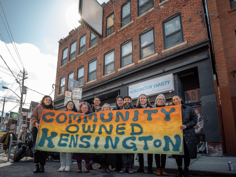 Members of the Kensington Market Land Trust stand in front of 54-56 Kensington Ave. in Toronto holding a multicoloured banner reading Community Owned Kensington. (Supplied photo/Kensington Market Land Trust)