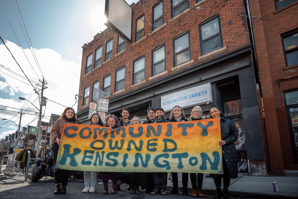Members of the Kensington Market Land Trust stand in front of 54-56 Kensington Ave. in Toronto holding a multicoloured banner reading Community Owned Kensington. (Supplied photo/Kensington Market Land Trust)