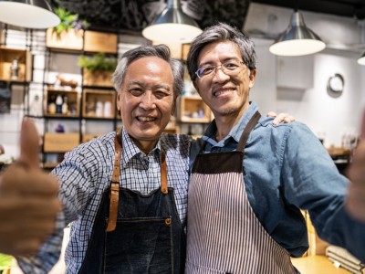 Two men wearing aprons look into the camera while giving the thumbs up sign in a grocery store.